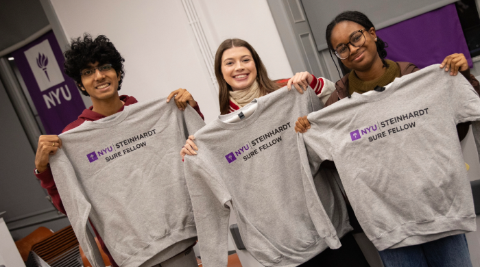Three students hold up sweatshirts that say NYU Steinhardt SURE Fellow 