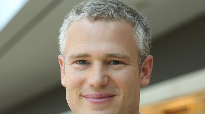 Headshot of Matthew Kraft wearing a dark suit jacket, and a blue collared shirt, with a slight smile against a softly blurred background.