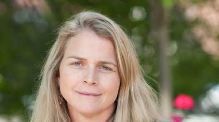 Headshot of Meredith Rowe wearing a short-sleeved red top and smiling slightly, with a softly blurred outdoor background.
