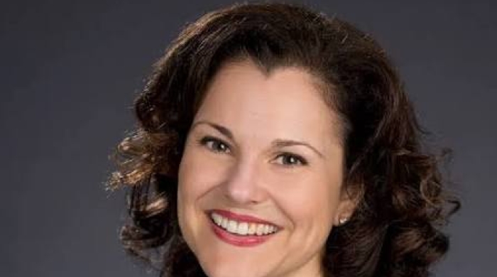 Headshot of Rebecca Callahan smiling, wearing a navy blazer, dark top, and gold necklace against a dark gray background.