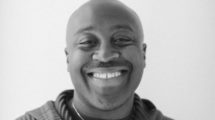 Black and white image of a man wearing a hoodie and necklace, smiling in the center of the frame