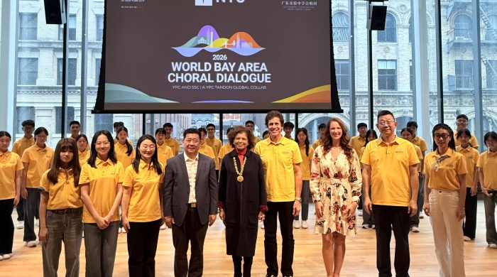 A photo of two rows of people in a large room smile for the photo in front of a projected slide with logos and the text "2026 World Bay Area Choral Dialogue"