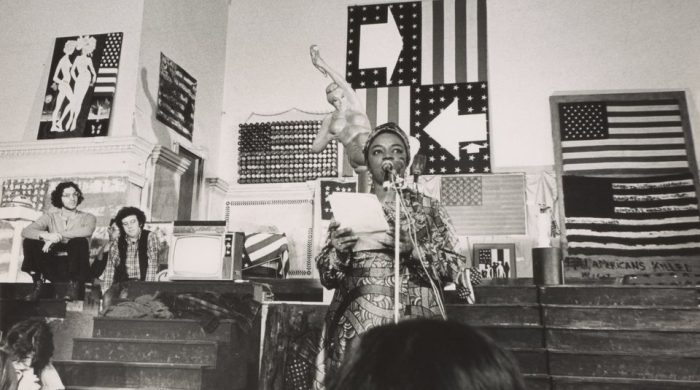 Black and white image of Faith Ringgold wearing a patterned dress as she speaks into a microphone while holding a piece of paper.