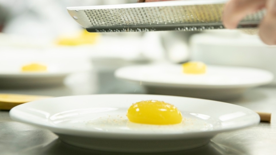 A student holding a grater over an "egg" made out of mango, yogurt, milk, and agar.