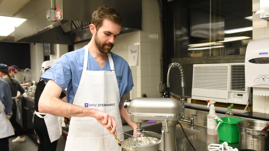 A student in scrubs and an NYU Steinhardt apron scrapes the inside of a stand mixture with a spatula.