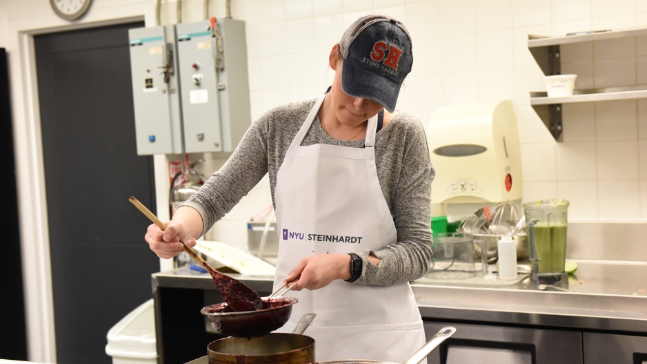 A student stirring a berry mixture through a sieve wearing an NYU Steinhardt apron.