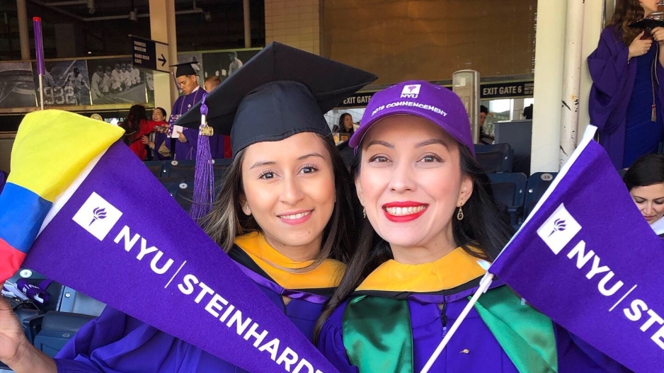 Two CSD alumni in graduation gowns holding pennants that say NYU Steinhardt.