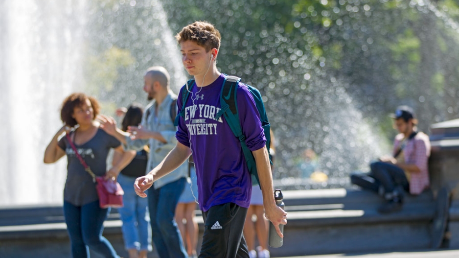 An undergrad student wearing an NYU T shirt walks past Washington square fountain