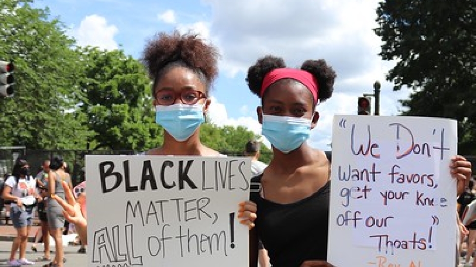 Two young black girls hold protest signs reading "Black Lives Matter, All of them!" and "'We Don't want favors, get your knee off our throats!' - Rev Al."