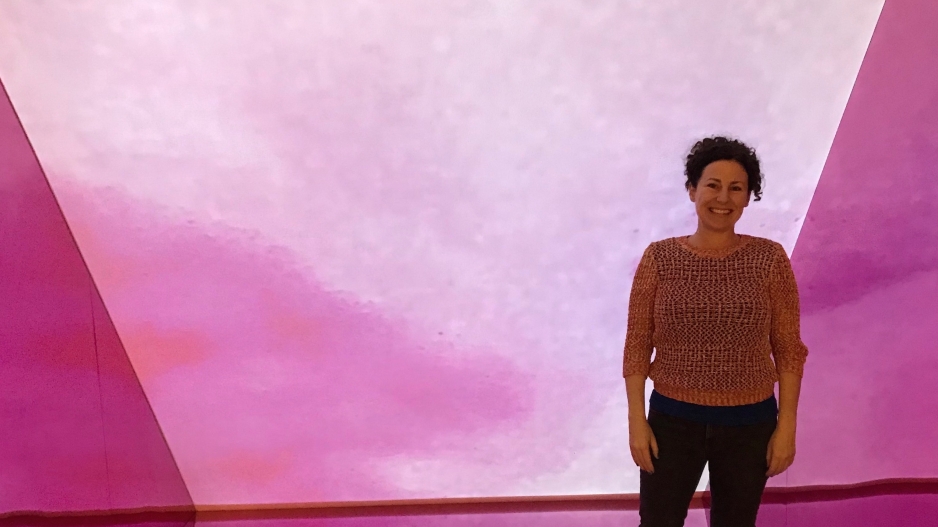 Mara Mills, a middle aged white woman with brown curly hair in a messy bun, stands in what looks like a pink box or stage set, the artwork we are all Pink Inside by Christen Clifford