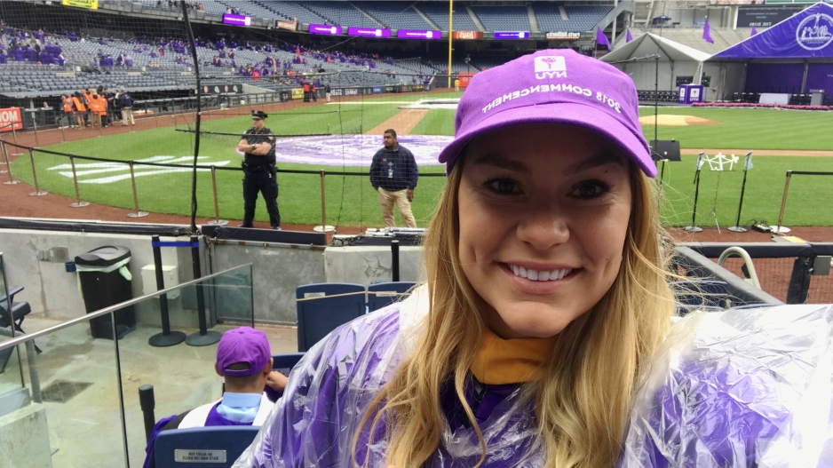 Jessie Warren wearing a purple NYU baseball cap and rain poncho at graduation at Yankee Stadium.