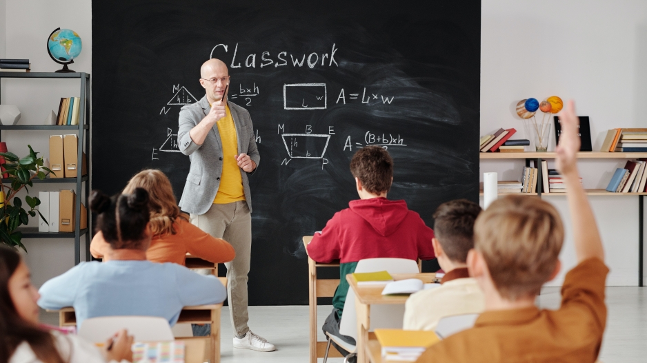 Teacher Stands at the front of the room pointing to a student with their hand raised