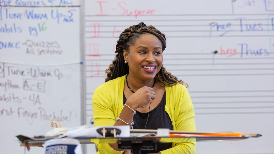 A music instructor who is a Black woman sits at the front of the room, smiling, with a violin on her lap