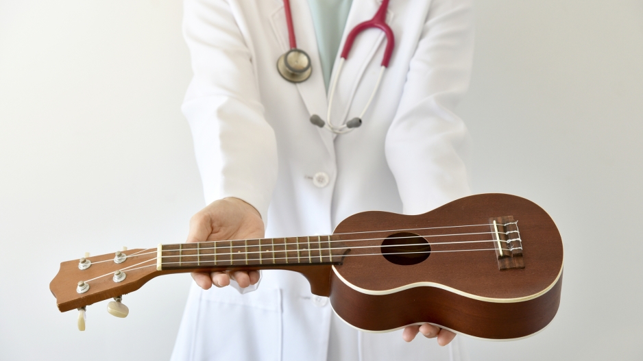 A doctor in a lab coat with a stethoscope around their neck holding out a ukulele with extended arms