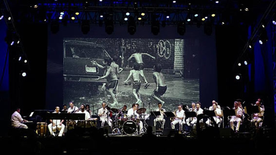The Multiverse Big Band performing on stage in front of a screen showing a B&W film of young boys in the street playing in the water from a fire hydrant