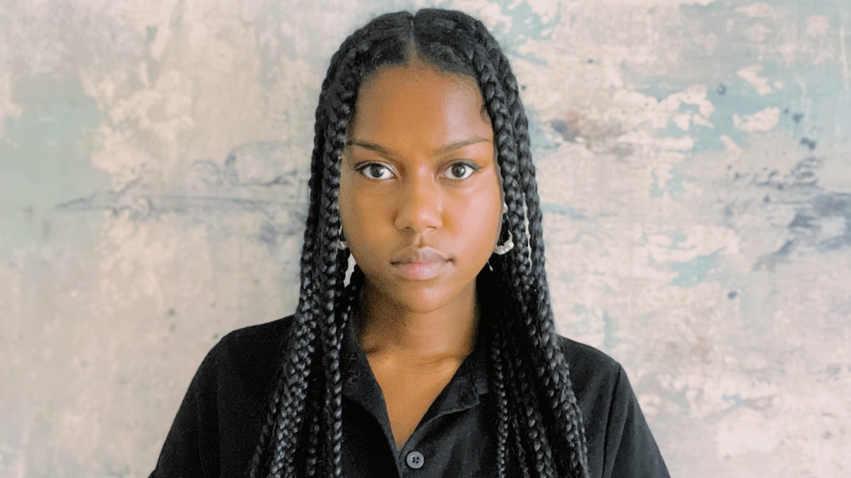 An image of Maya Beverly, wearing a short-sleeve blouse and looking directly into the camera. She is against a bluish background.