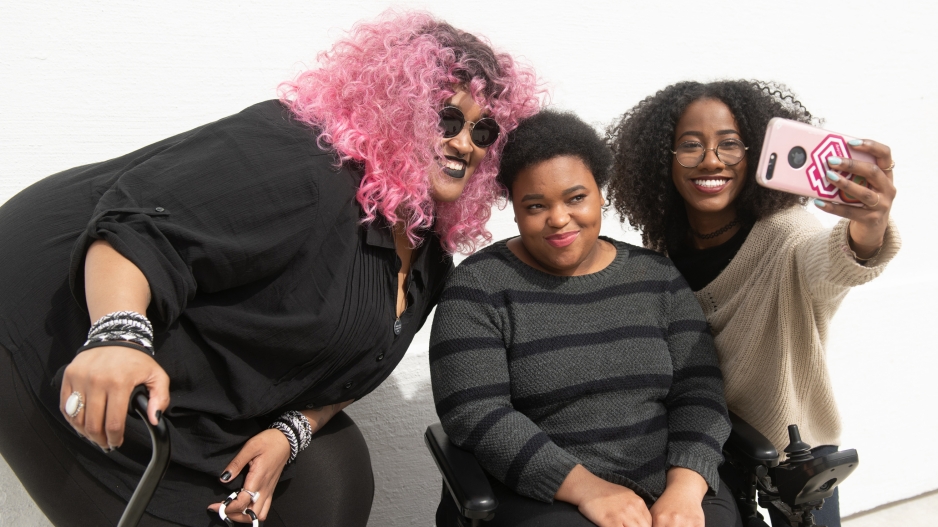 A tight crop of three Black and disabled friends (a non-binary person with a cane and tangle stim toy, a non-binary person sitting in a power wheelchair, and an invisibly disabled femme) smiling and taking a cell phone selfie together.