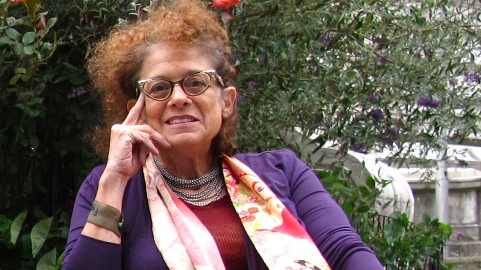 Simi Linton, a white woman, with reddish hair and black cats eye glasses, is seated in a power wheelchair in a garden. She is wearing a purple jacket, beaded metallic necklace, and long multi-colored scarf.  She is smiling.