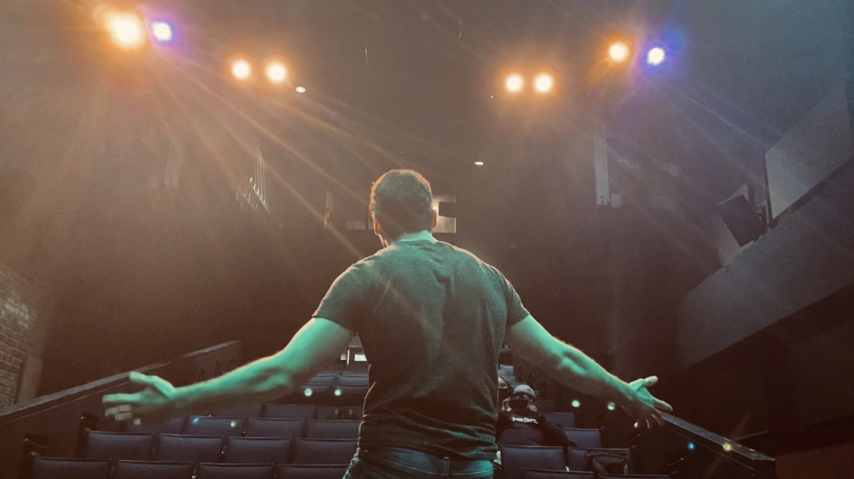 The back of a performer with outstretched arms facing the audience in a mostly empty theatre shot from the back of the stage