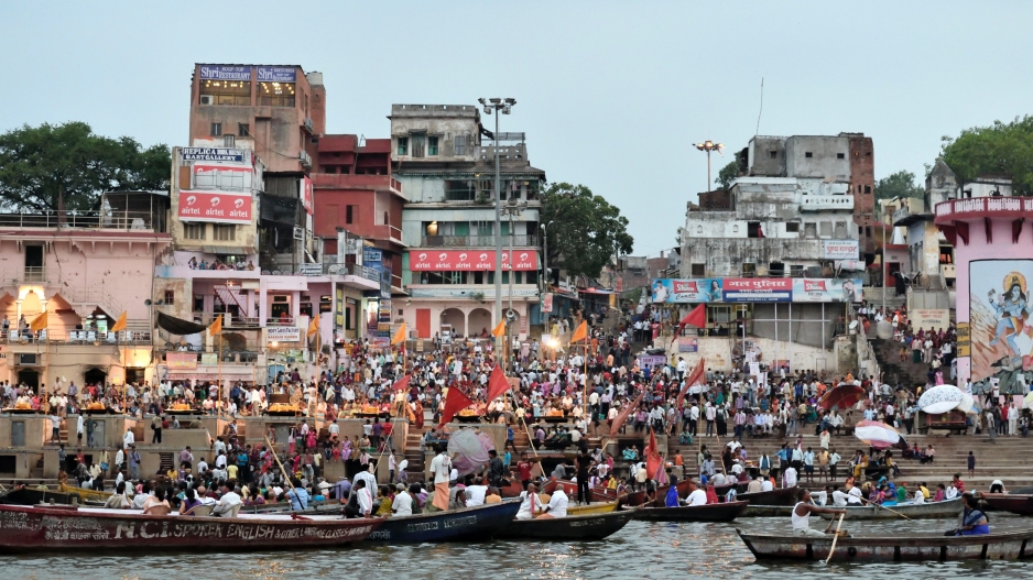 A photo of a skyline of Uttar Pradesh, India taken from the water with boats in front. There are many people crowded around the shore.