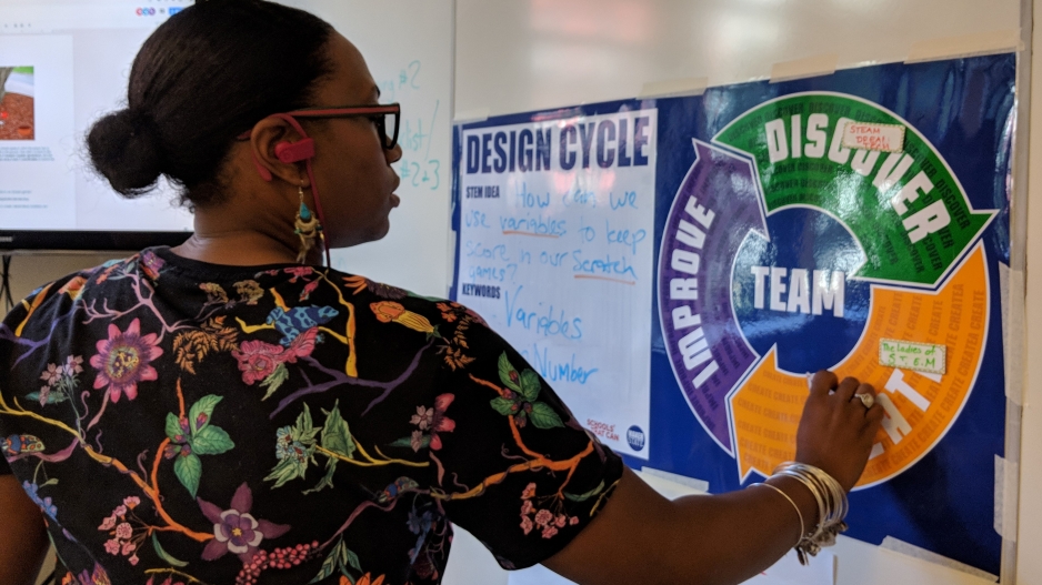 A female teacher stands in front of a white board and writes on a blue poster of the MPP design cycle.
