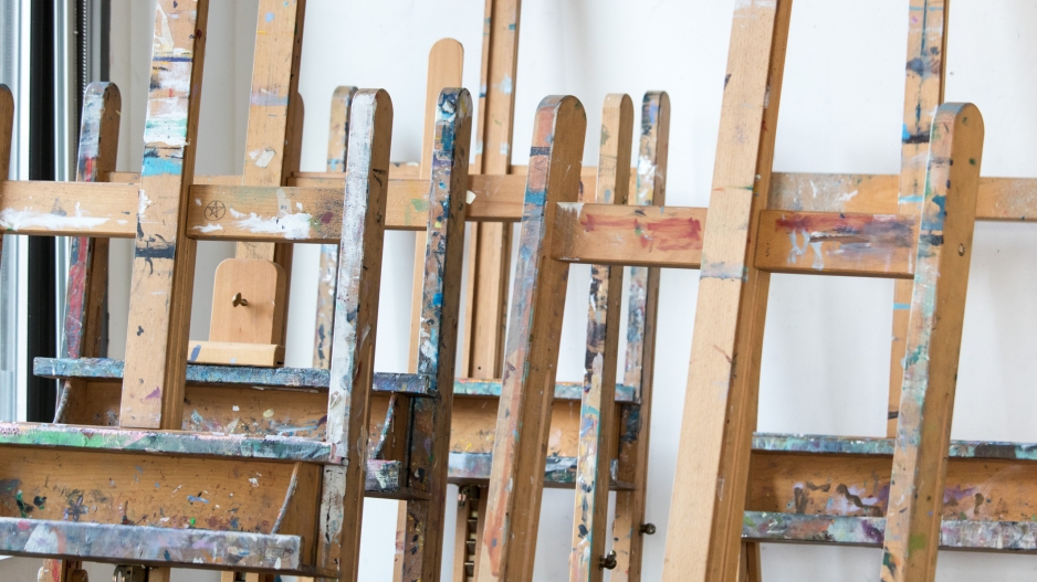A closeup image of easels in a painting studio. The easels are made of light wood with colorful paint splatters. The wall behind them is white.