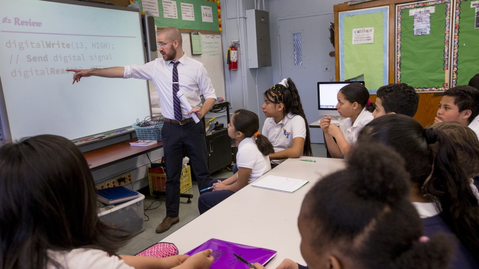 A teacher is standing at the front of a classroom filled with students in uniforms. He is pointing at the screen, which is showing a review of the content.