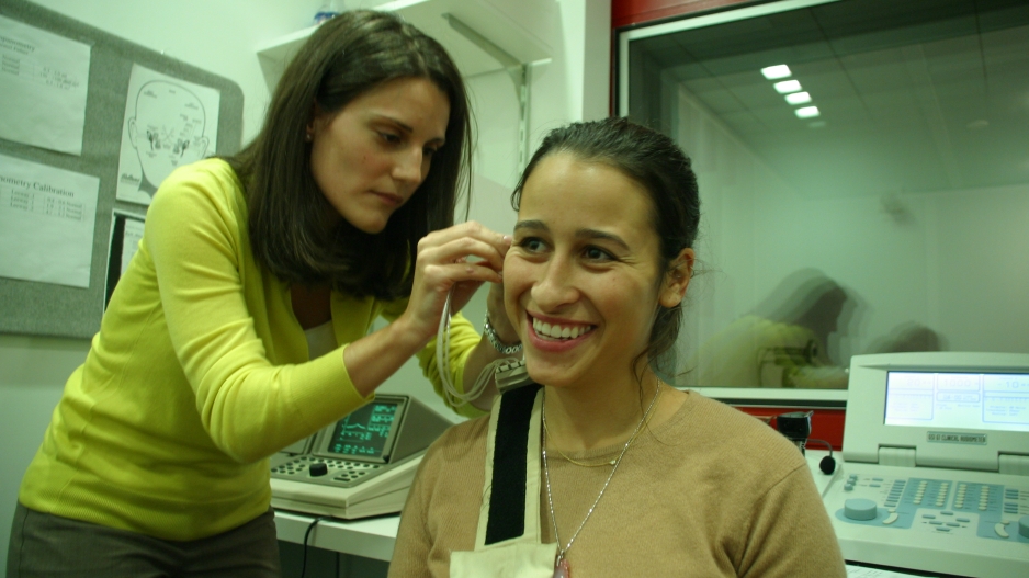 One woman is having a device connected to her ear by another woman. The first woman is smiling.