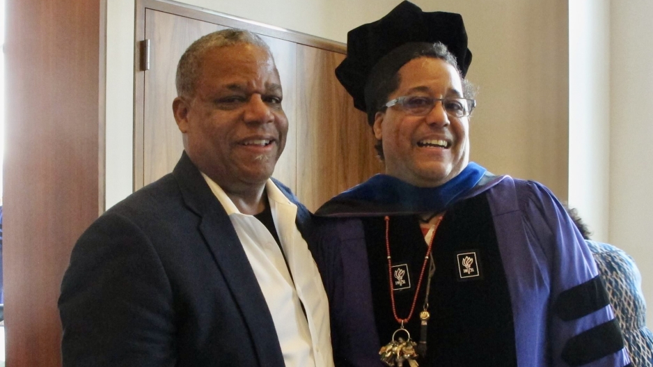 Two men, one in a white shirt and navy blue jacket, and the other wearing a doctoral robe stand in front of a door.