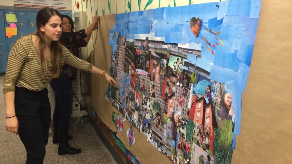 A young woman points to a banner created by her students. Another woman hangs a corner of the banner on a school wall. The banner is a collage with buildings, pictures of students, and a bright blue sky.