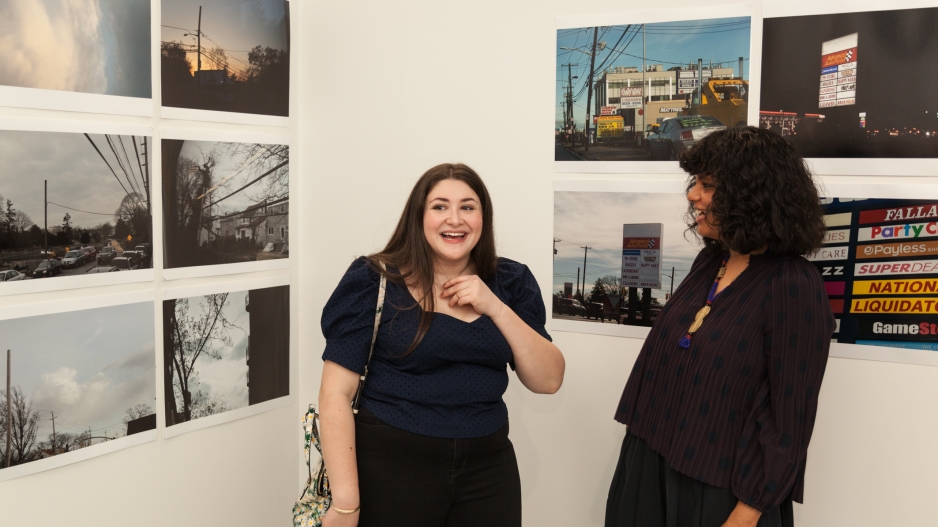 Two art department students appear in front of a white wall with photographs pinned to it in a grid. The students are talking and laughing.