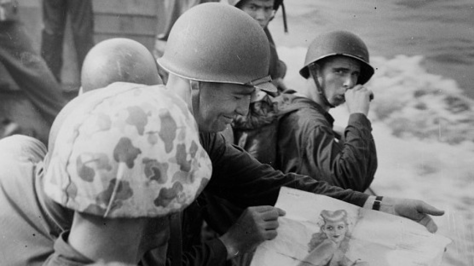 A black and white photo of four soldiers. There are four helmets pictured in the photo. One soldier is holding a picture of a pin up.