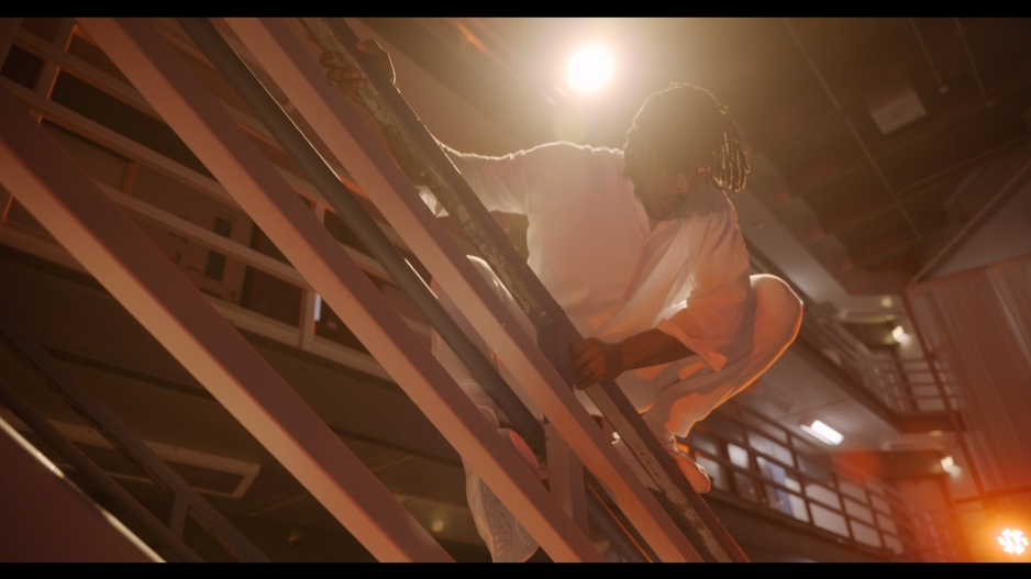 A Black man in a white uniform climbs on the metal railing of prison stairs.