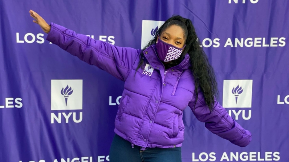 Arianna Deans, a Black woman in a purple coat, stands in front of an NYU-branded backdrop.