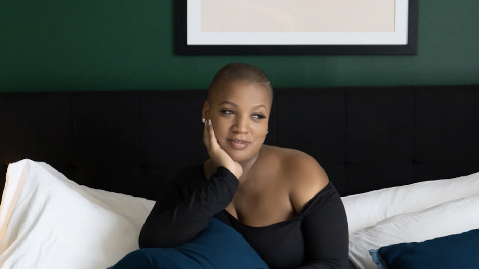 Jade Kearney, a Black woman with short hair, sits on a bed looking contemplatively to the side. A framed print behind her reads "New York" in several fonts. 