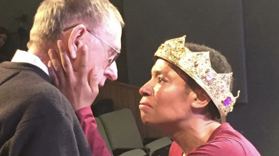 A Black woman, wearing a gold crown, cradles the face of an older white man as they face each other. 