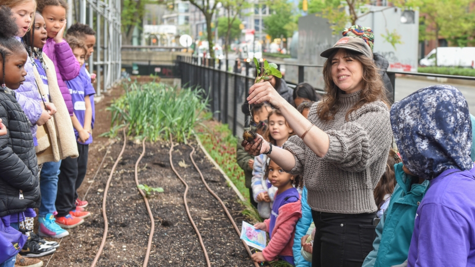 A volunteer from NYU's Urban Farm Lab stands next to a seed bed, explaining something to a group of children.