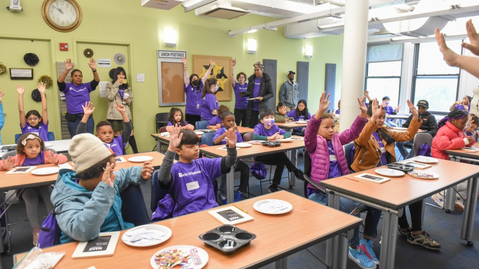 Children in an art classroom raise both hands. On the tables are art supplies.