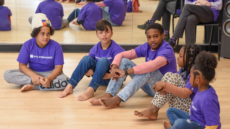 Children, dressed in purple NYU Steinhardt shirts, sit in a line in a dance studio.