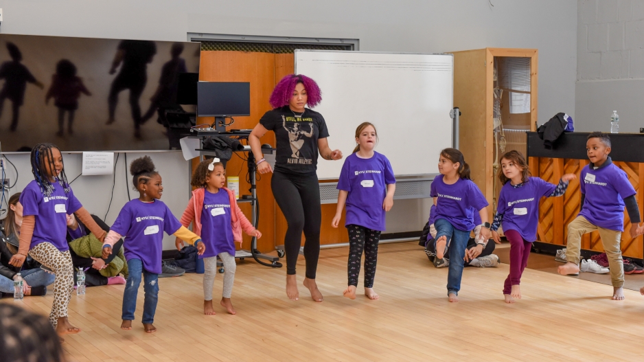 Katherine Jimenez, a woman with magenta hair, demonstrates a dance move to a group of children who imitate her by standing on one foot. 