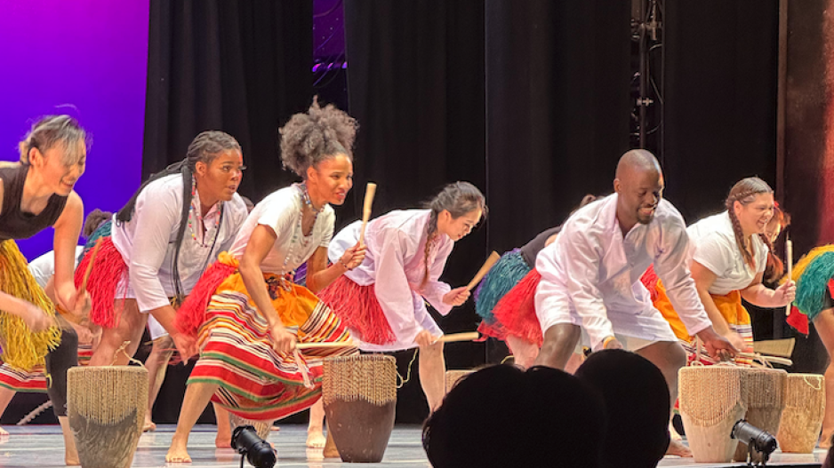 A group of drummers, dressed in colorful skirts, perform on stage.