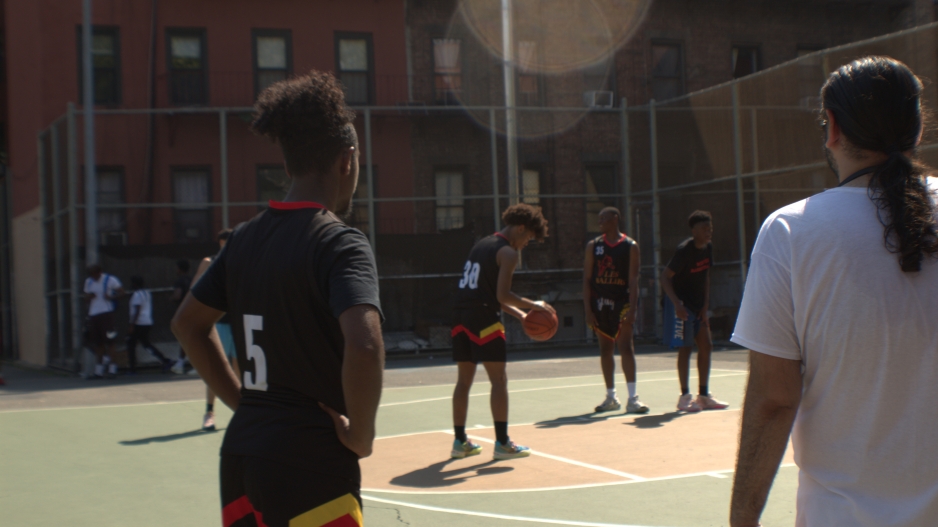 photograph of five players playing basketball in an outdoor court, four players are in black uniforms and one of the players is dribbling and preparing for a free throw. 