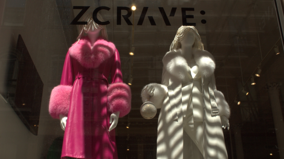 Picture of two plastic mannequins in a storefront, one is wearing a pink fur robe and the other is wearing the same design of robe in white and holding a beaded sphere purse. The text “ZCRAVE:” is placed on the window in between the viewer and the mannequins.
