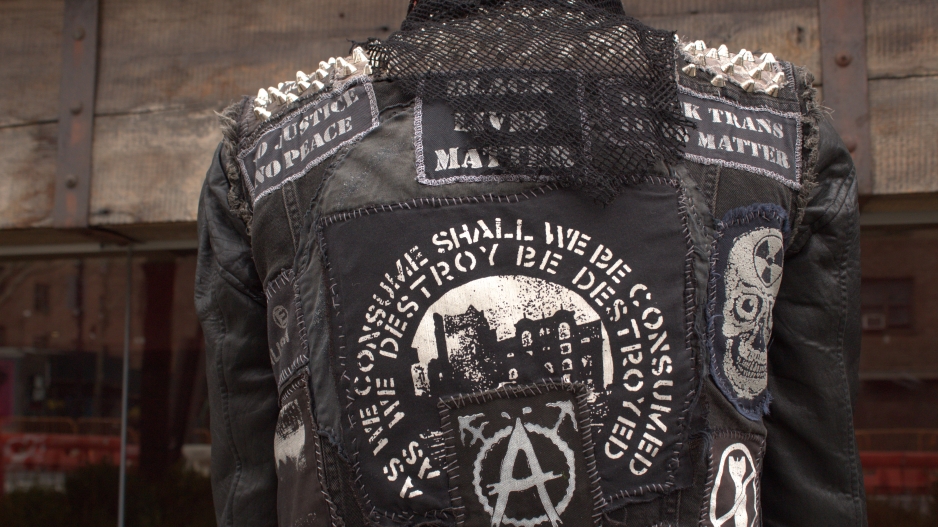 Photograph of the back of someone wearing a designed black jacket with many patches of graphic text sewn on, including “BLACK TRANS LIVES MATTER” and “BLACK LIVES MATTER” and a skull patch. 