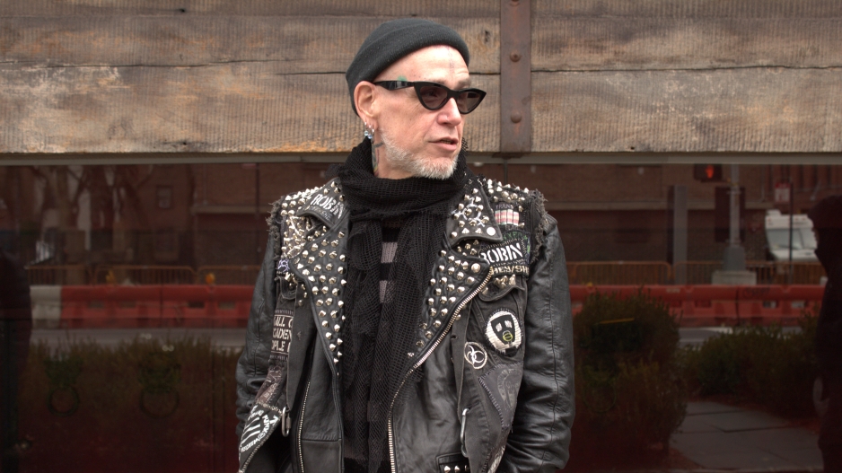 Photograph of the front of an older white man wearing black sunglasses, a black beanie, and a black leather jacket with sequins and patches. The man is looking to his left with his mouth slightly open. 