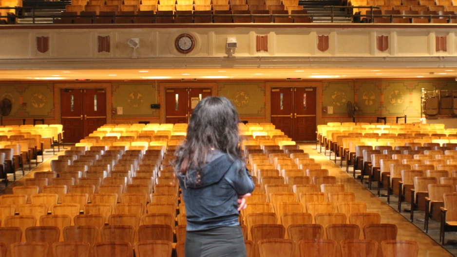 Photograph of the back of a brown haired girl standing on a stage in front of a large empty audience of wooden chairs in an auditorium. 