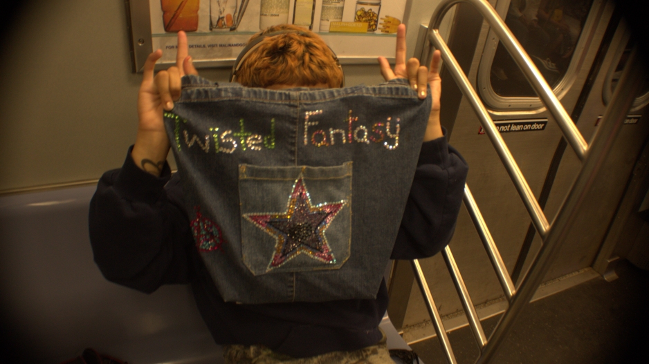 A person from the waist up with orange short hair holding up a bag that has “Twisted Fantasy” written out in gems and a star on the pocket. The person is holding up rock on signs with both hands and is sitting in the subway with army pants on.