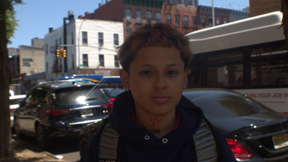 Photograph of a young boy with eyebrow slits and orange hair wearing a snazy sweatshirt and backpack. The boy has small hoop earrings and a serious face. 