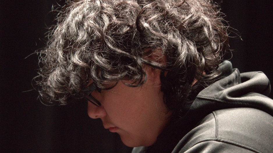 Side profile of a young boy with curly black hair wearing a black hoodie with a pitch black ground. 