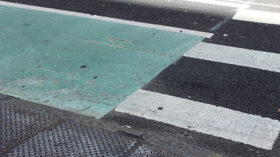 Photo captures the black and white lines that make up a cross walk at the end of street  corner. A portion of the cross walk in covered in blueish colored, plastic tile